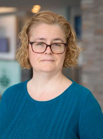A photo of a white woman staring directly towards the camera. She has light, curly hair and is wearing a blue shirt. She is wearing glasses and has a stoic look on her face.