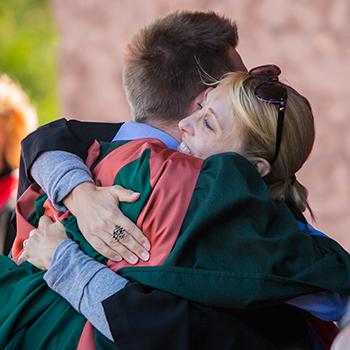 A male student in convocation gown hugging a professor on the Bata podium in the summer sun