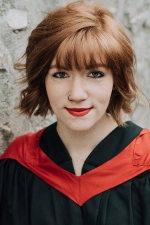 Emilee Storfie smiling at the camera, wearing a convocation gown and red hood, on a stone background