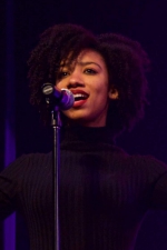 Mikeela Skellekie singing behind a microphone stand, against a black backdrop