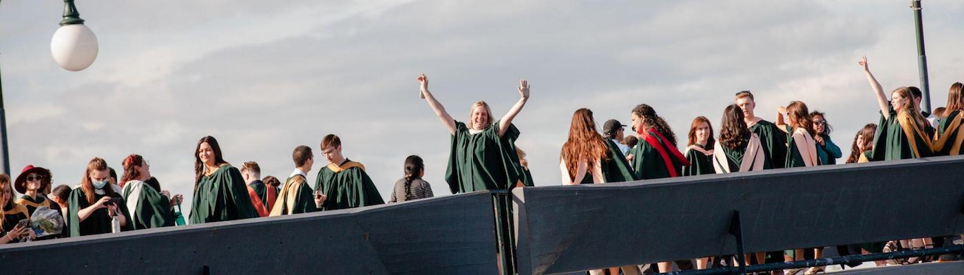 Image of many grads in their regalia on the Faryon Bridge. Woman in the centre is smiling at the camera with her arms in the air.
