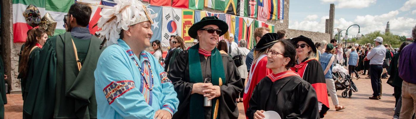 Image of three faculty wearing Convocation regalia and traditional Indigenous regalia. They are smiling at each other.