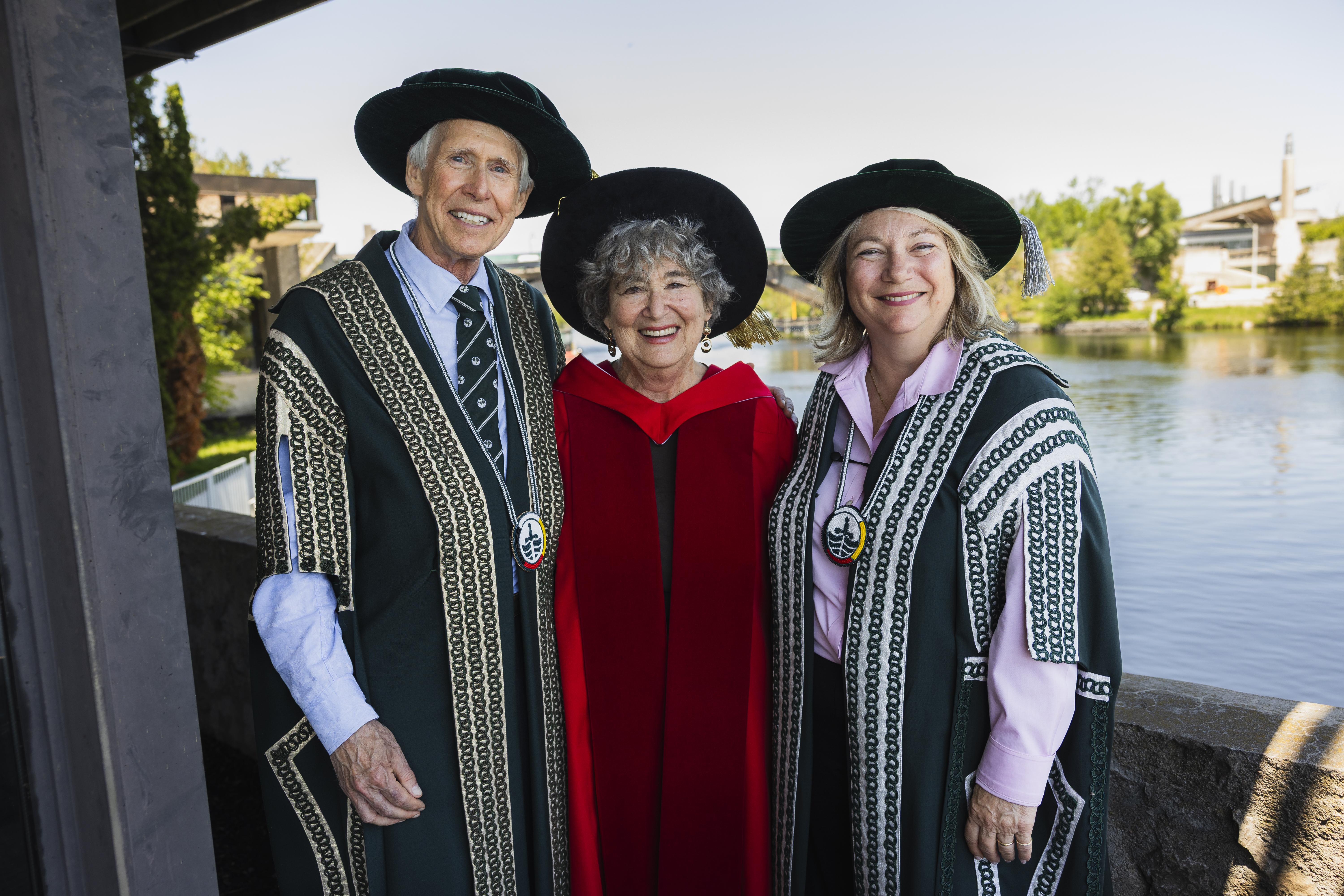 Three people standing outside in regalia, smiling at the camera