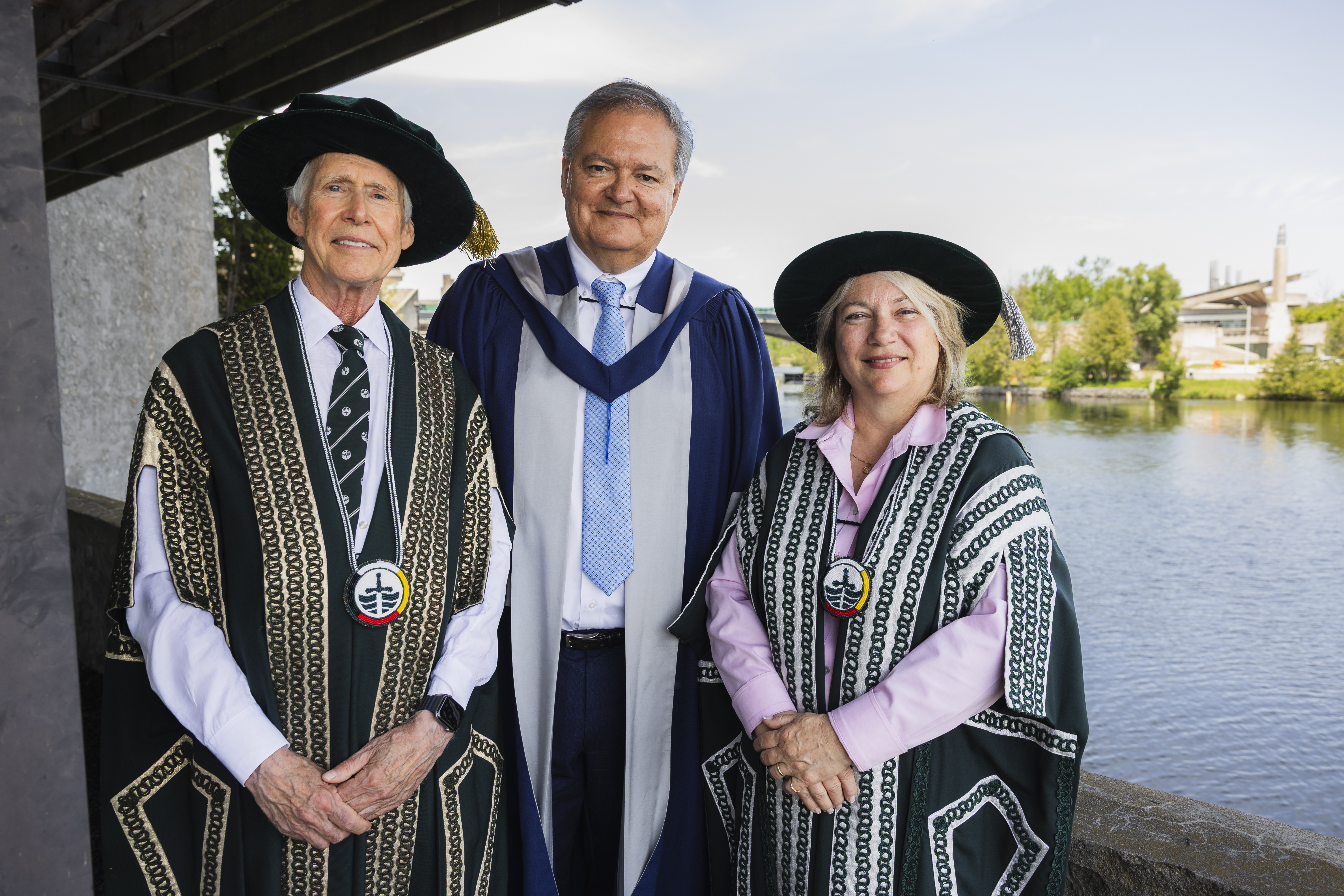 Three people standing outside with academic regalia on,the river behind them