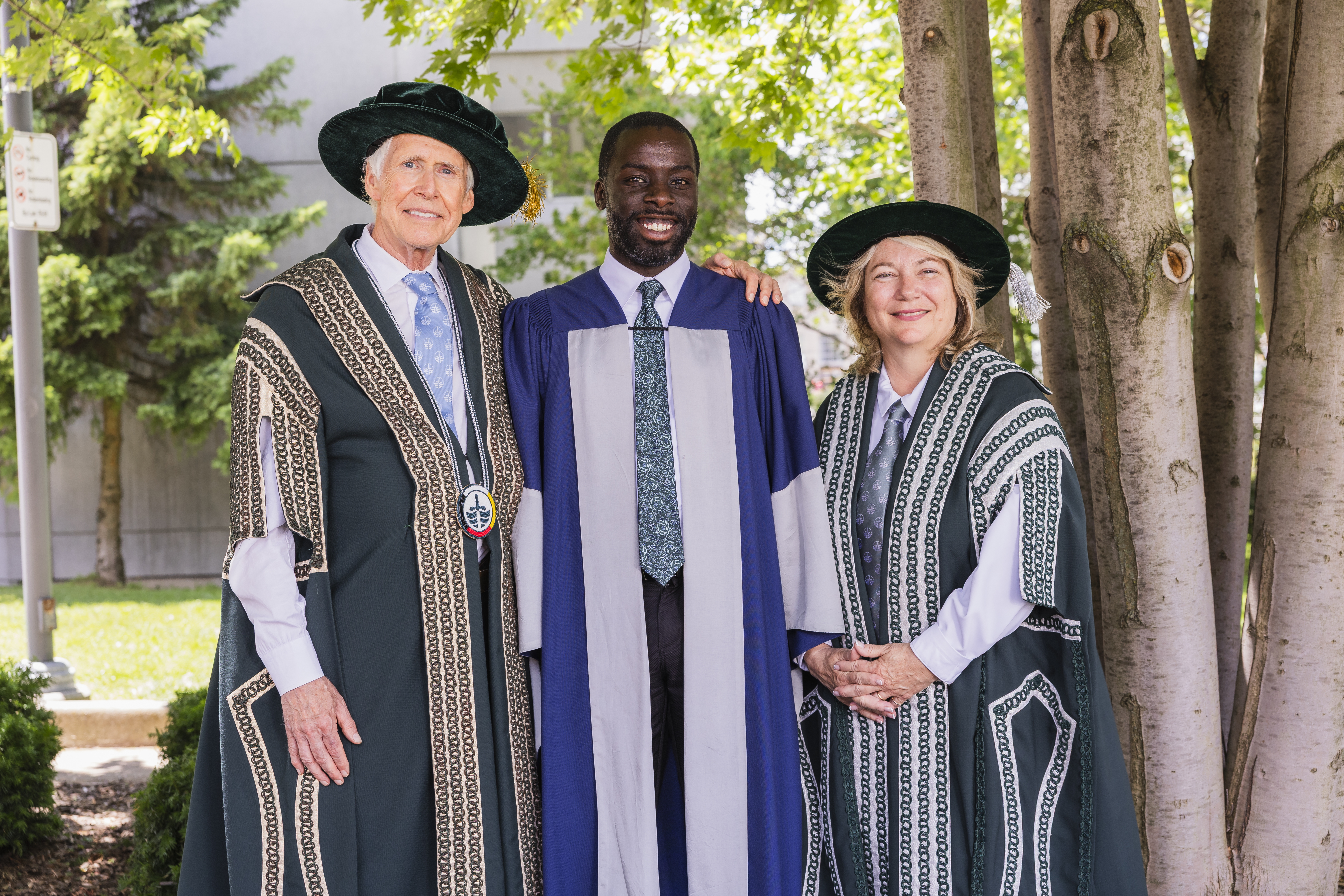 Three people standing outside underneath trees dressed in academic convocation regalia