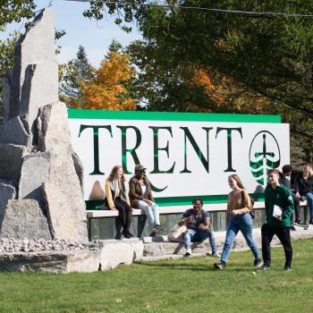 Trent University Peterborough Campus sign with students lounging around. 