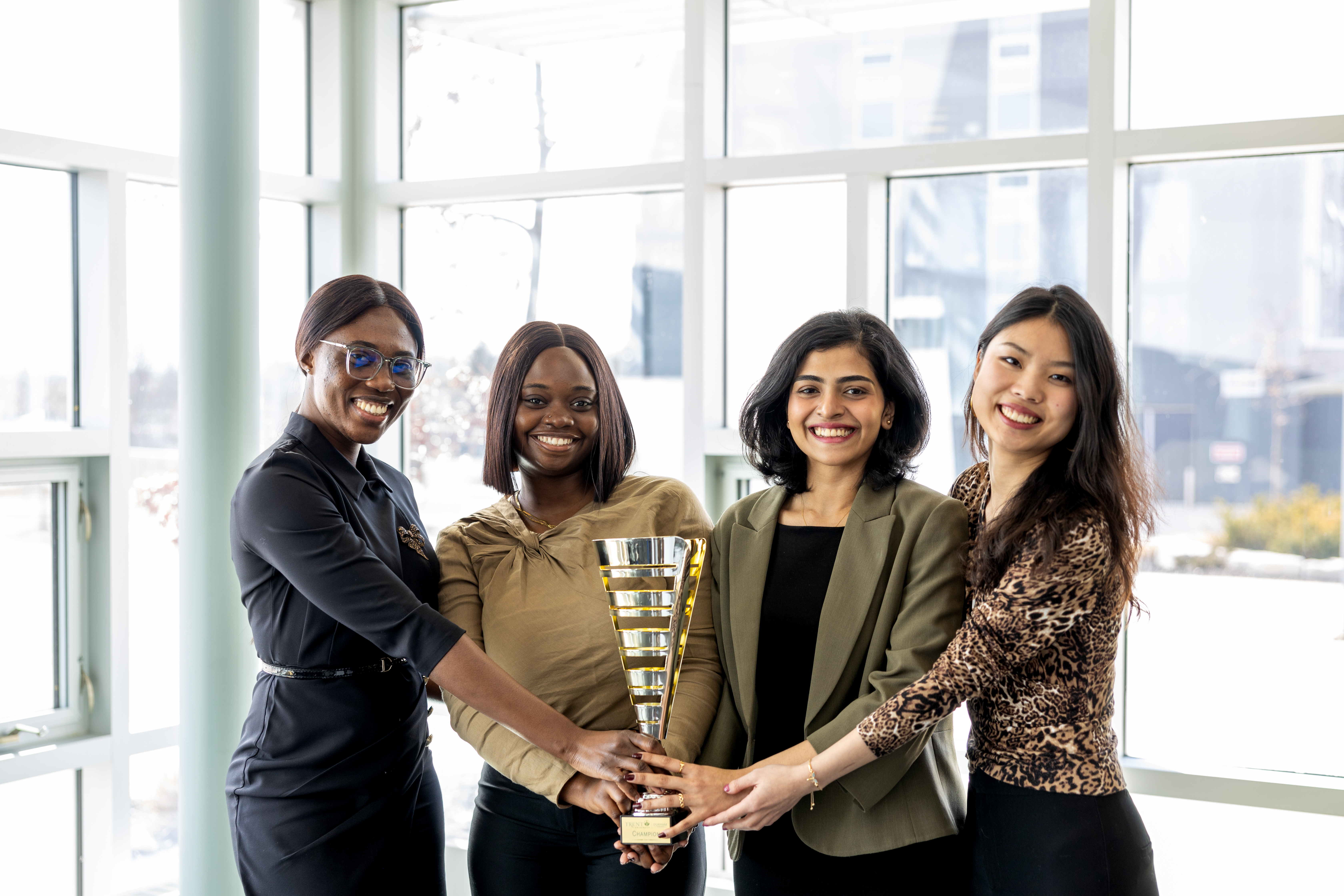 Four women smiling and holding a gold trophy together in a bright, window-lit space.