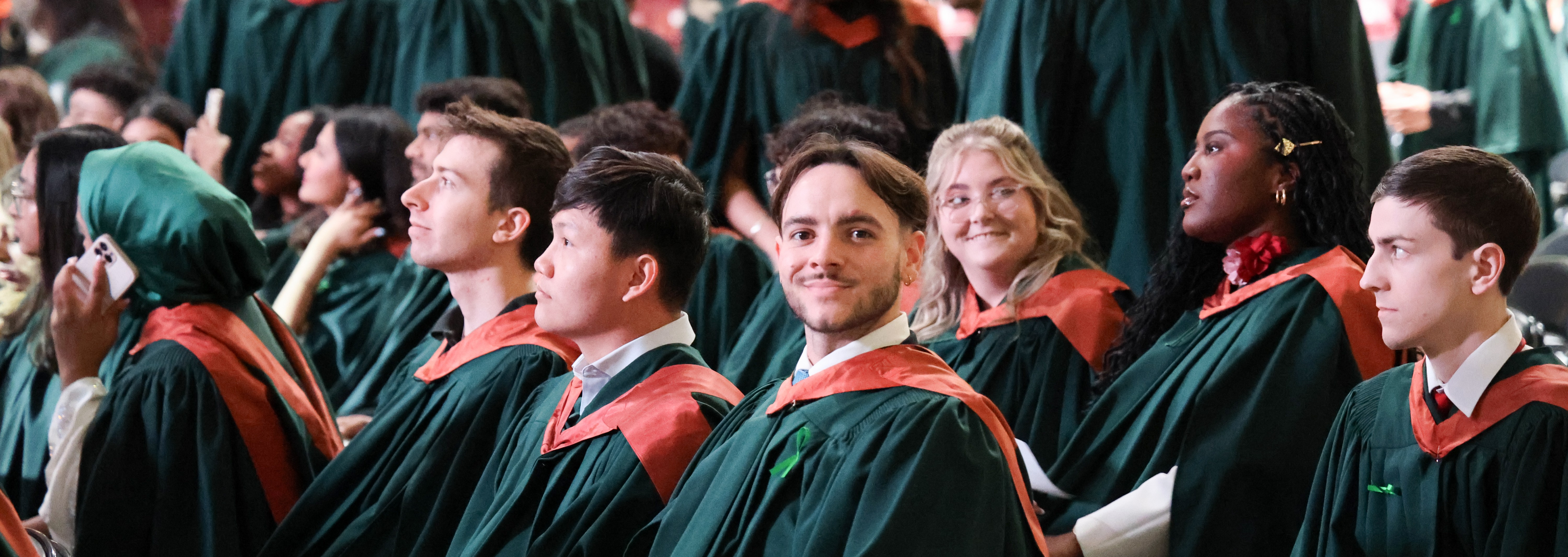 Graduates in green gowns sit in an auditorium during a ceremony, with one smiling student looking toward the camera.