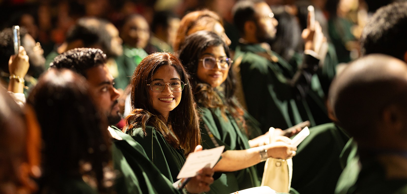 Graduates in green gowns sit in an auditorium during a ceremony, with one smiling student looking toward the camera.