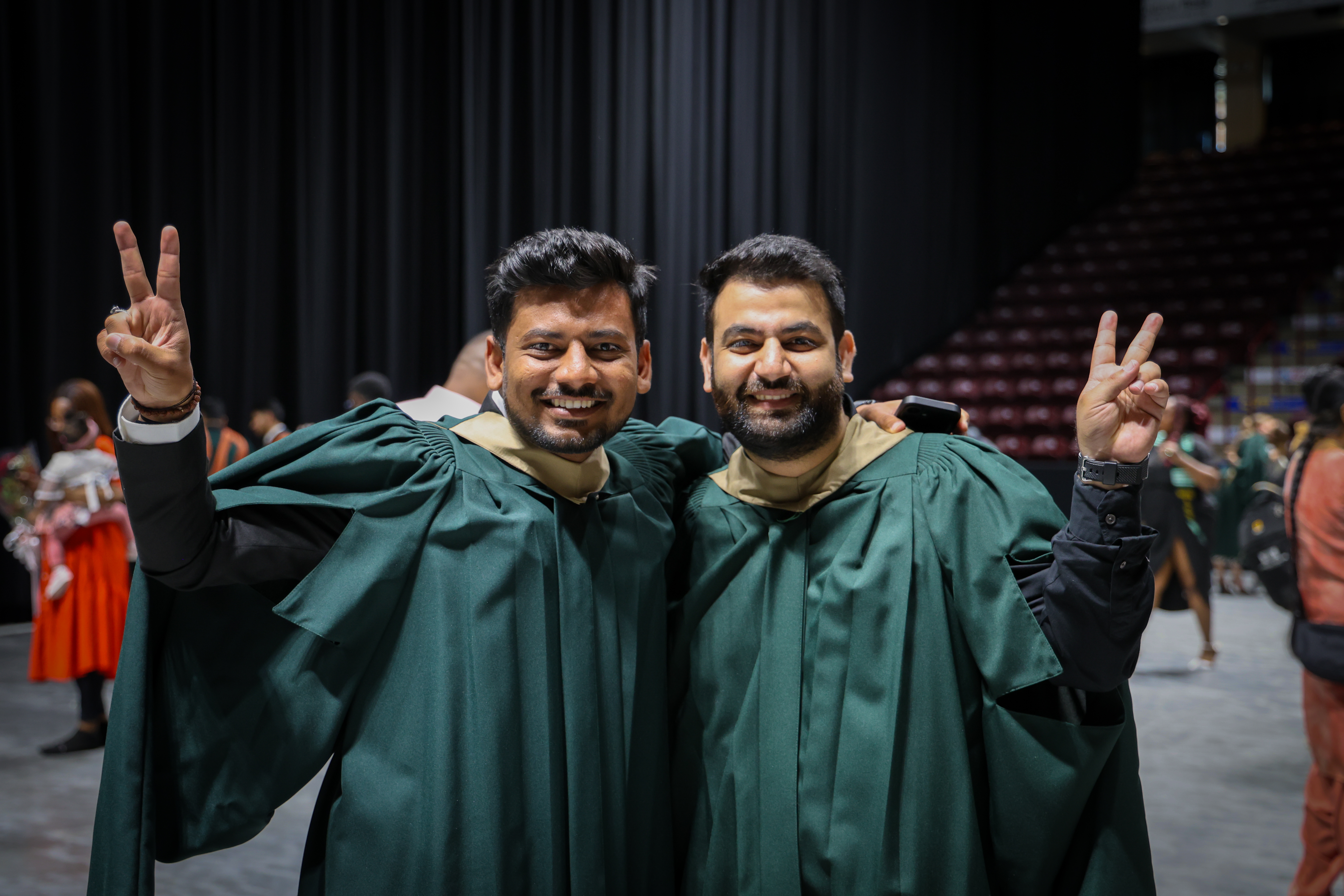Two graduates in gowns smile and pose together indoors, holding up peace signs during a convocation celebration.