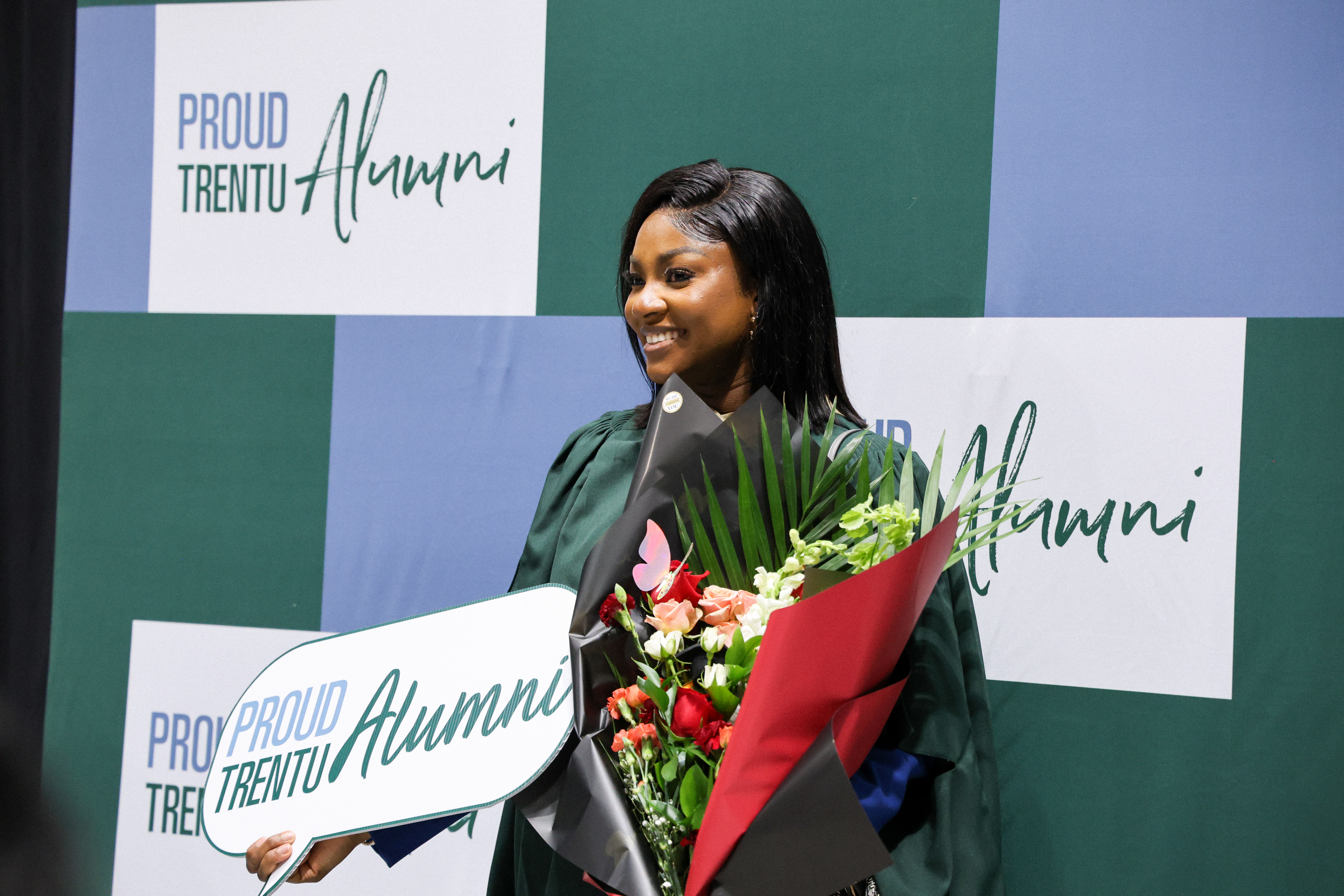 Graduate in a gown smiles while holding a bouquet of flowers and a “Proud TrentU Alumni” sign in front of a photo backdrop.