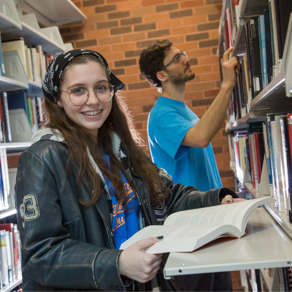 Young woman with an open book looking at the camera and man check out a section in the library behind her
