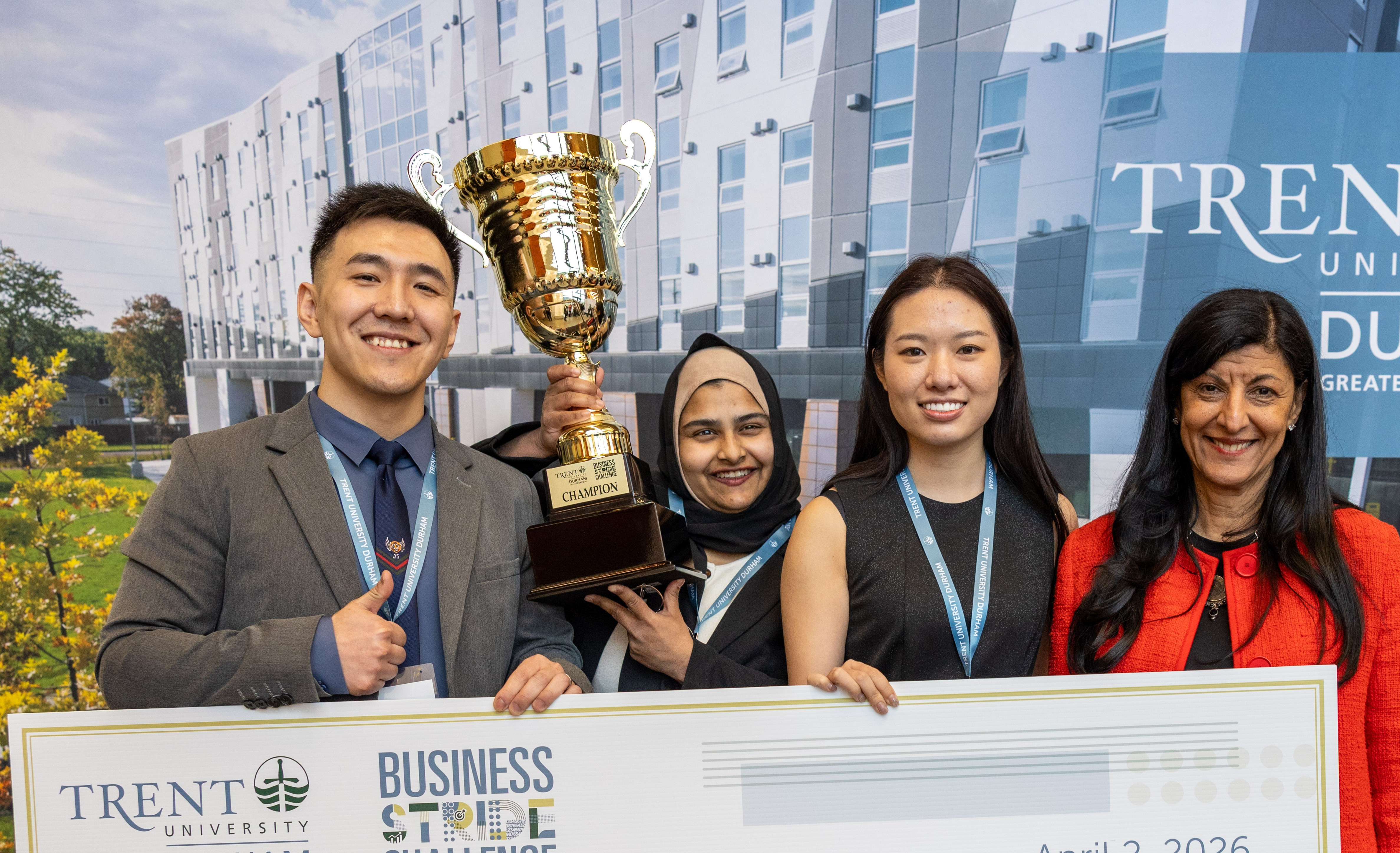 Three students and an instructor pose with a trophy and a large $1,500 cheque for the Trent Durham Business Stride Challenge. The cheque is dated April 2, 2026, and names the winning team, T.Member. The group stands in front of a Trent University Durham GTA backdrop.