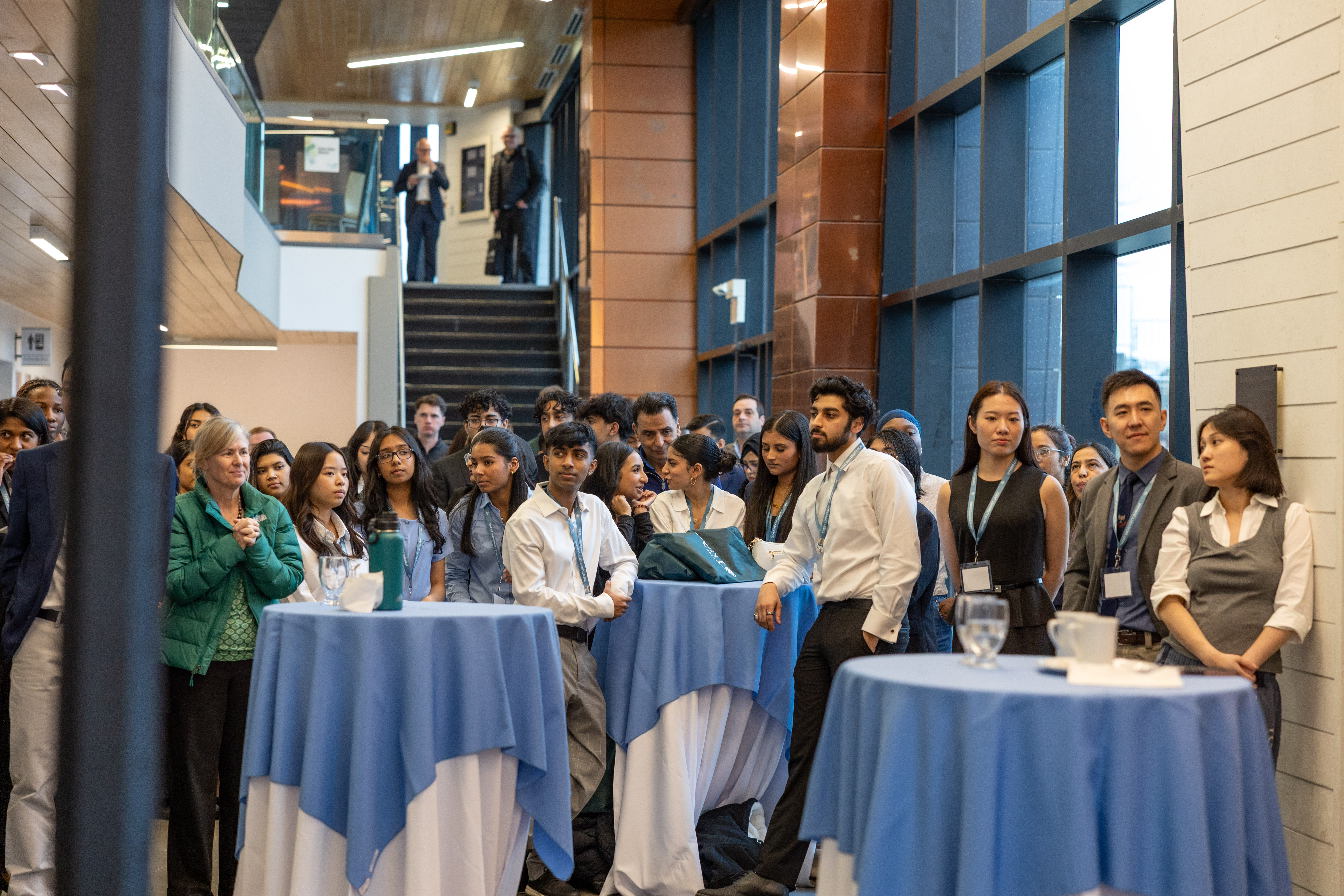 Students, staff and guests gather around cocktail tables in a bright campus atrium during the Trent Durham Business Stride Challenge. Several participants wear event lanyards, and two people stand on the staircase behind the group.