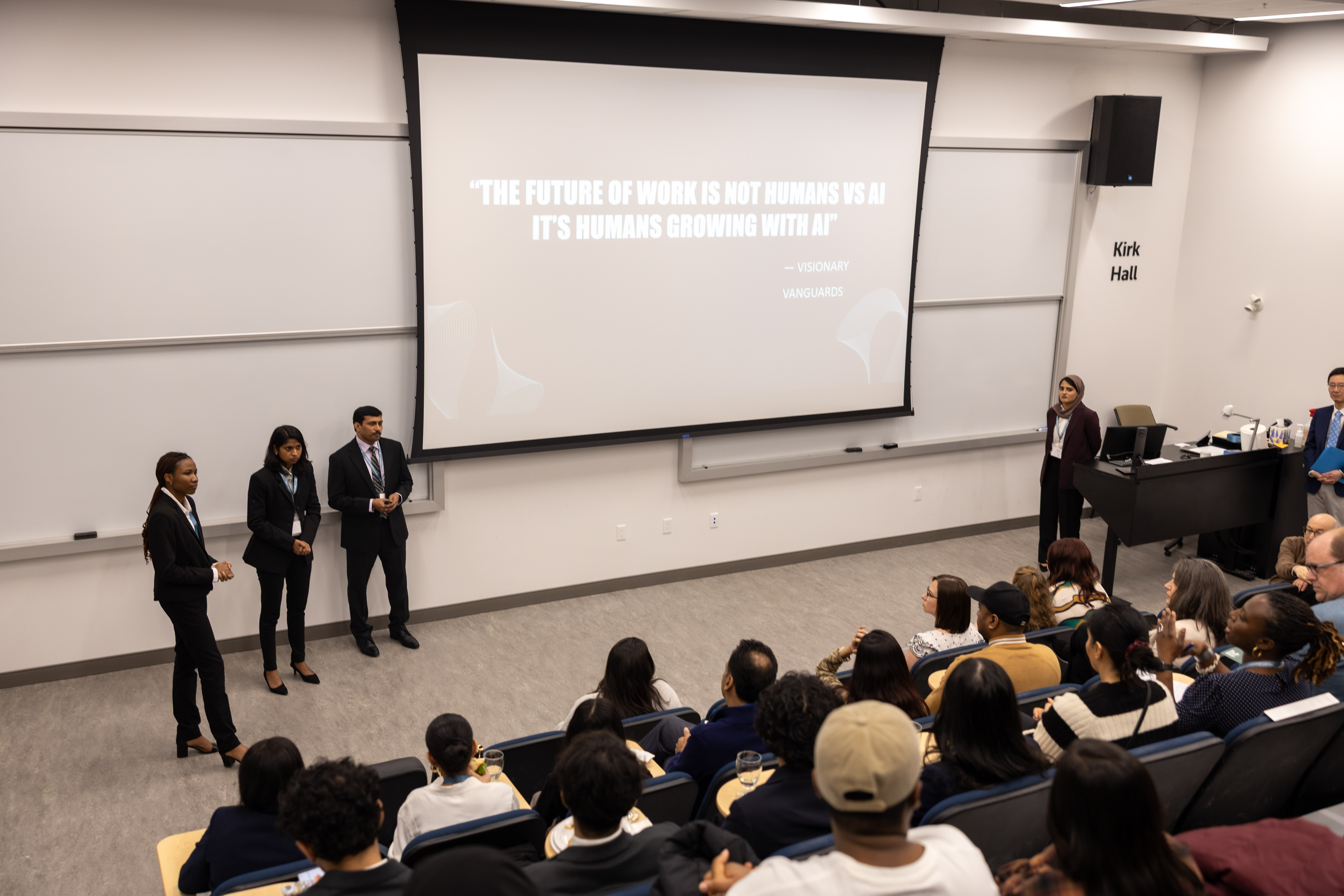 Three students present to an audience in a lecture hall. The screen reads, “The future of work is not humans vs AI; it’s humans growing with AI,” with attribution to Visionary Vanguards.