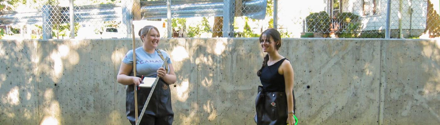 2 feamle students standing in a river in waders infront of a concrete wall, smiling at the camera in the summer shade of some trees