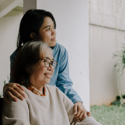 A candid indoor photo of two people near a window. One person is seated in the foreground wearing a light-colored sweater, while another person stands behind with a hand resting on the seated person's shoulder