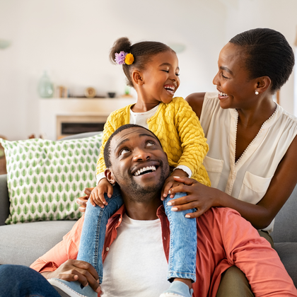 A family of three gathered in a living room. One parent is seated in the foreground while a child sits on their shoulders, and another parent sits nearby. The family is interacting together on a couch.