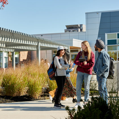 Three Trent University students wearing backpacks and carrying books stand together and talk on campus near a walkway.