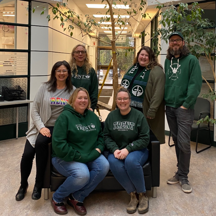 Group photo of Trent University Student Accessibility Services (SAS) staff standing together in Blackburn Hallway. Six staff members are gathered around a seating area, with some seated and others standing behind them. Several individuals are wearing Trent University apparel in green.