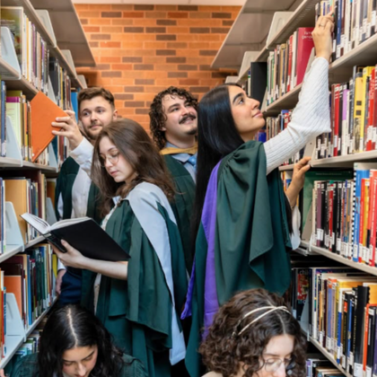 Copilot said:  Alt text:  A group of Trent Durham students studying and browsing books in the Trent Durham Library. Several students wearing green academic gowns are positioned between library shelves filled with books.