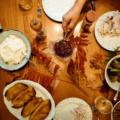 Overhead view of a dining table set for a shared meal. Dishes of mashed potatoes, cranberry sauce, sliced squash, vegetables, and carved turkey are arranged around the table, with autumn leaves used as decoration. A hand reaches toward the bowl of cranberry sauce in the center, surrounded by plates, utensils, and glasses, creating a warm harvest-themed gathering setting.