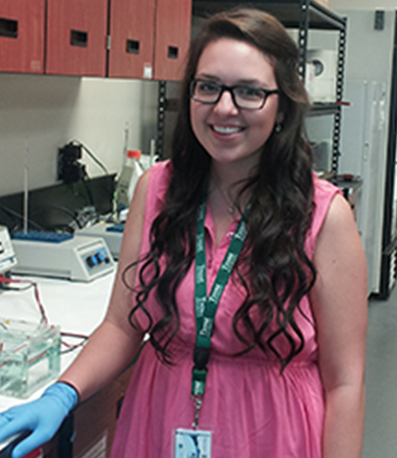 student Kristi Goulet standing in laboratory wearing pink dress and lab gloves smiling at camera