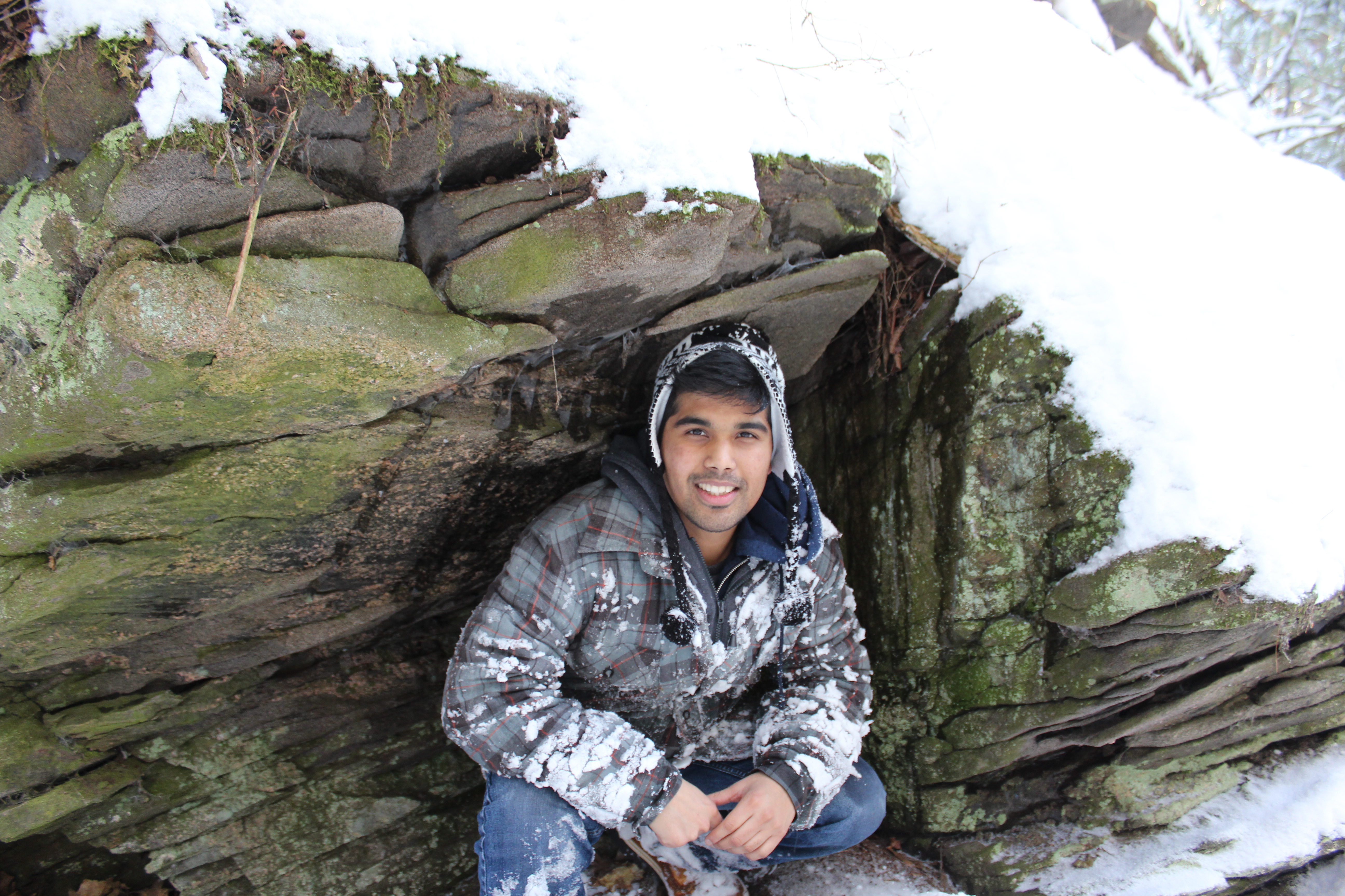 student, Vaughn Mengal, crouching under large rock near stream with snow around him 