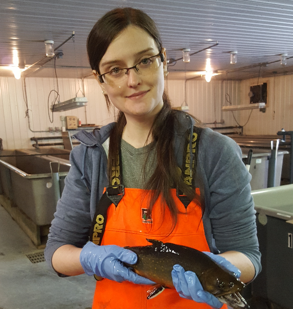 female grad student wearing bright orange coveralls holding an adult brook trout in a laboratory, smiling at camera