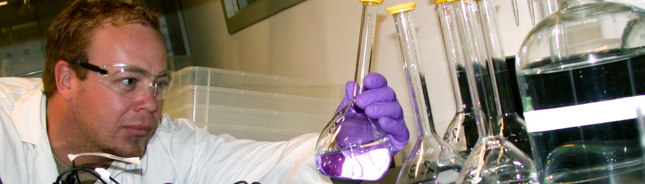 close up of male student wearing safety glasses, lab coat and gloves holding a flask and looking at it closely