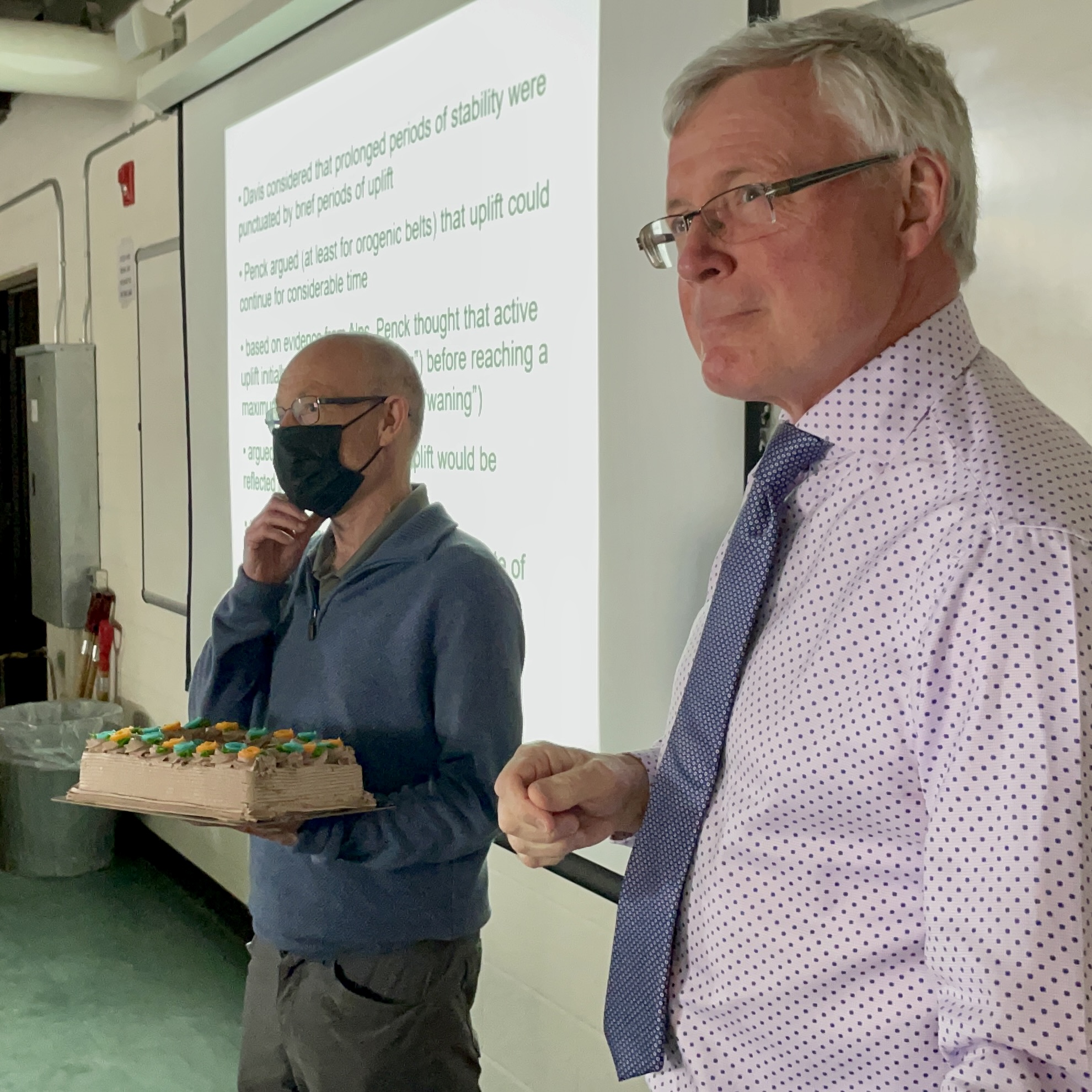 two white men in a classroom with a cake