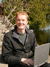 smiling man with pale skin and red hair outside on river with trees in background