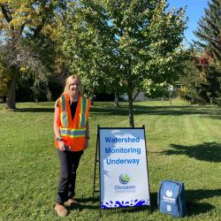 woman with blond hair and pale skin wearing bright orange safety fence in front of green trees and blue sky