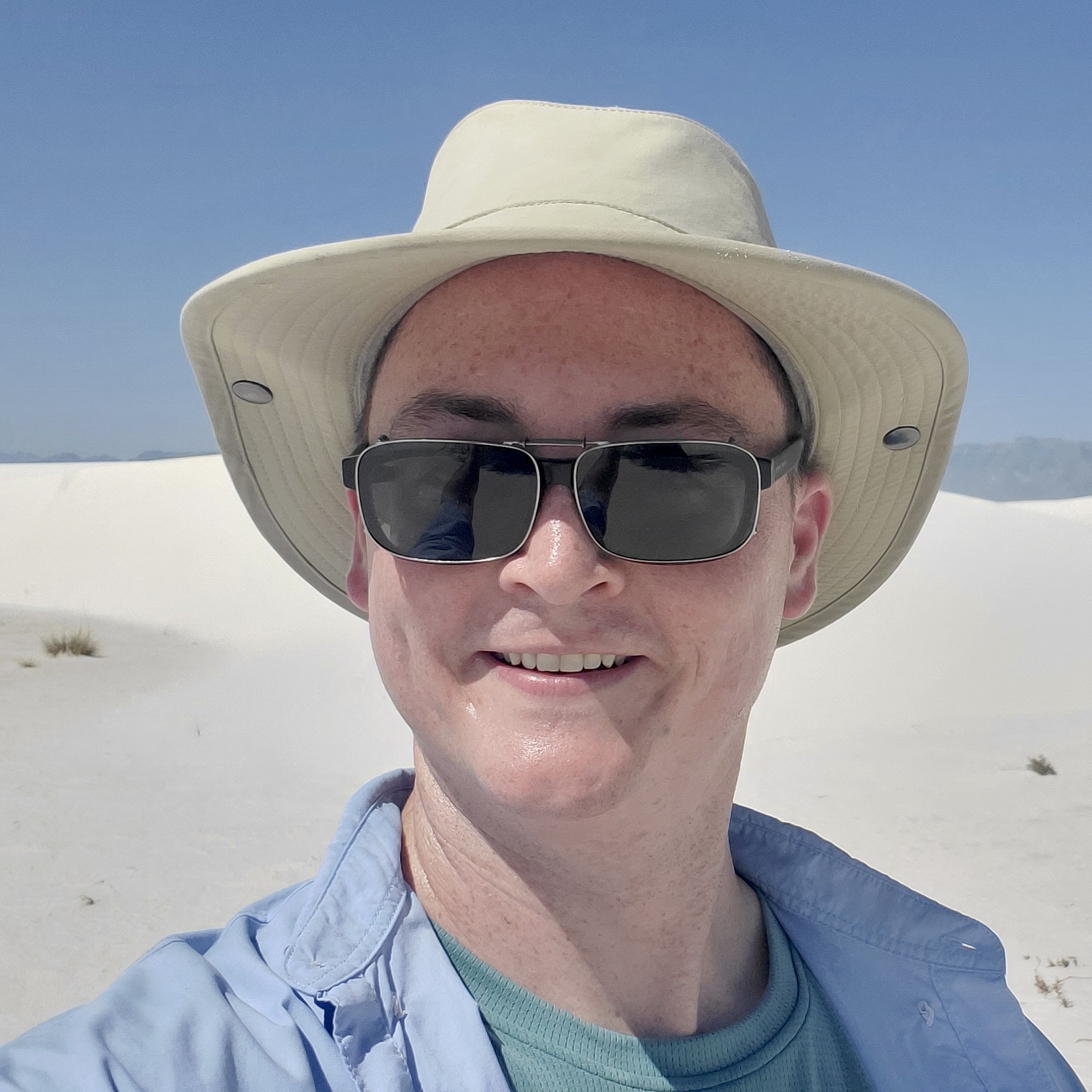 smiling person with pale skin wearing sunglasses, beige hat and blue shirt against a blue sky and sand background