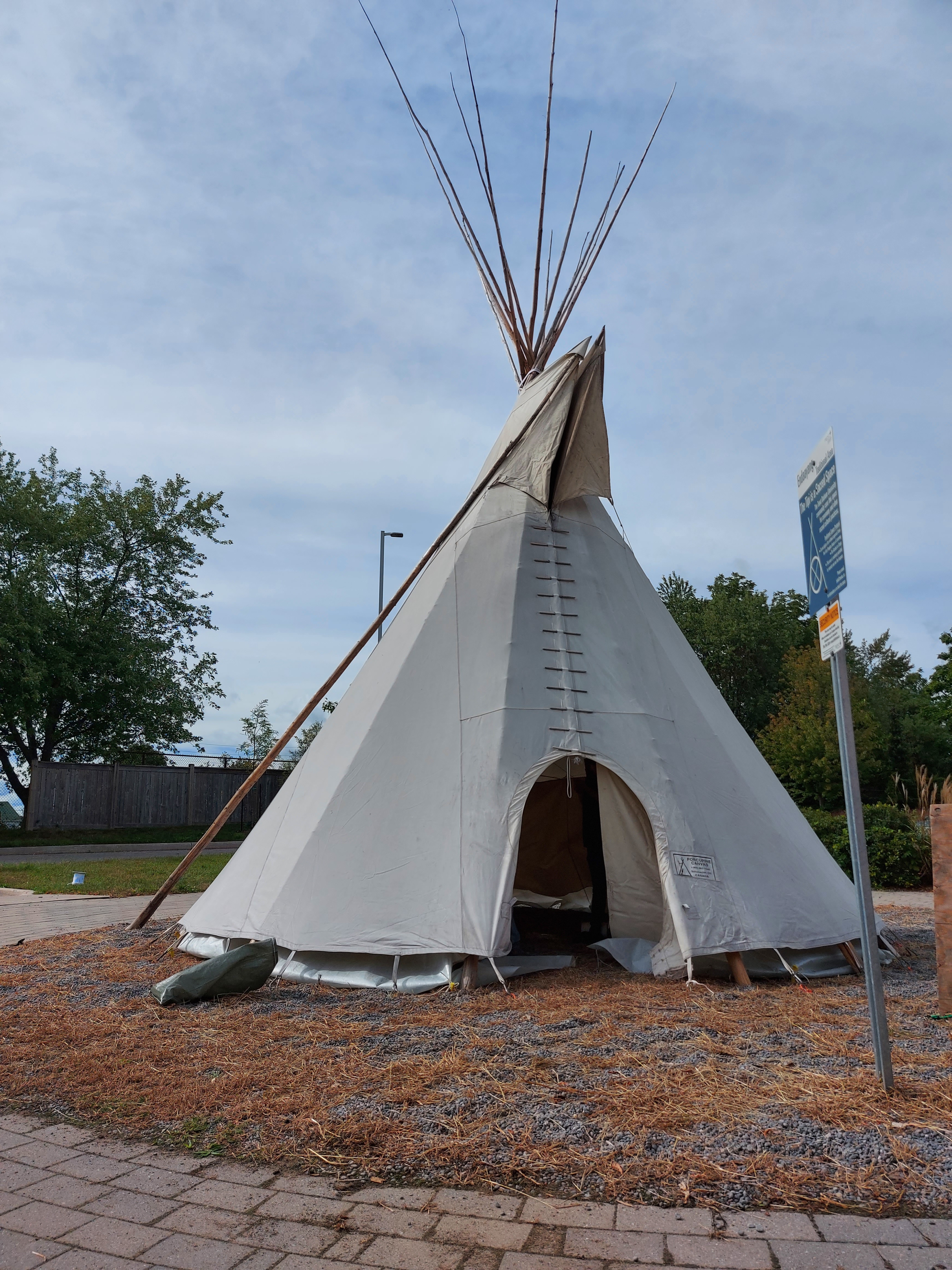A picture of the tipi located at the Endaayang traditional area. There is a bright blue sky behind it, and the tipi sits upon gravel terrain with a concrete walkpath nearby.