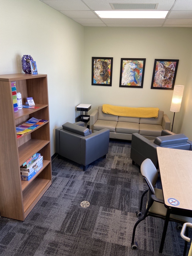 A picture of the Indigenous lounge. There is a couch, two chairs, a bookshelf with supplies, and several pieces of Indigenous artwork on the wall.