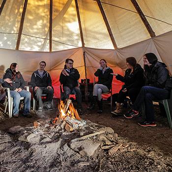 Students listening to a professor's lecture sitting in chairs inside of a teepee around a fire