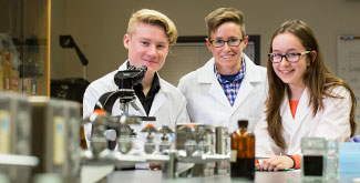 3 students in white labcoats, smiling at the camera, standing at a table behind a microscope and jars