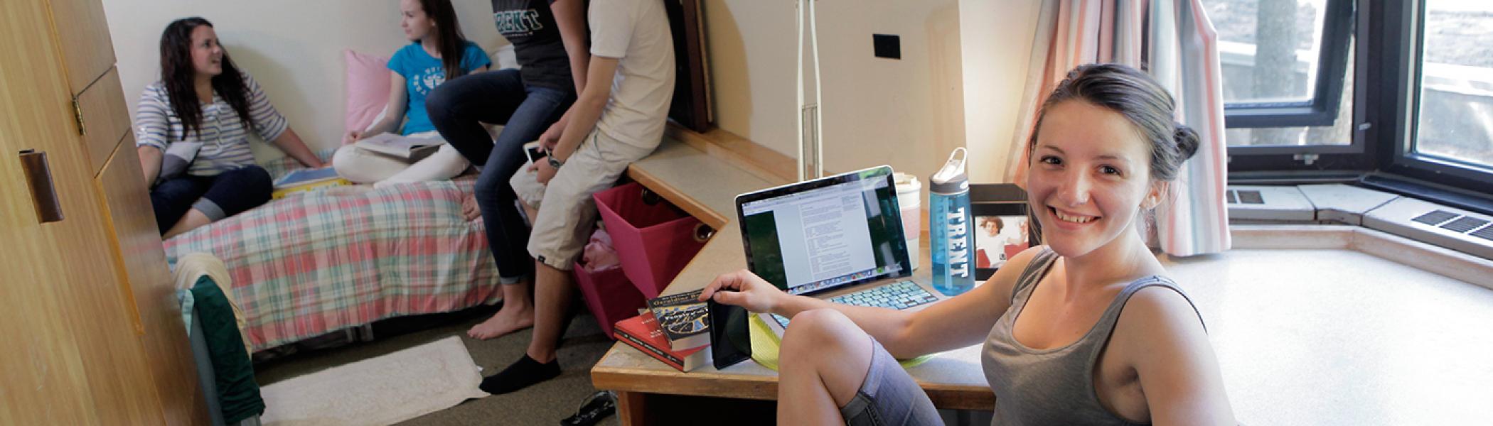 Student sitting at her desk in her Lady Eaton College residence room. Other students lounge on her bed in the background.