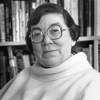 A black and white photo of Margaret Laurence, sitting in front of some bookshelves looking at the camera