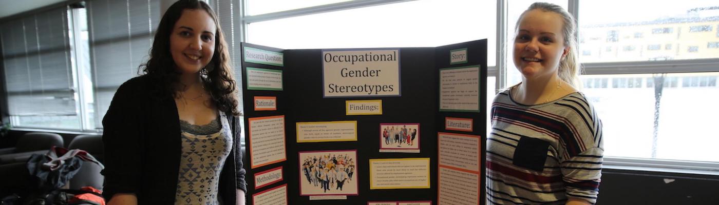 Two girls standing in front of a poster with the title &quot;Occupational Gender Stereotypes&quot;