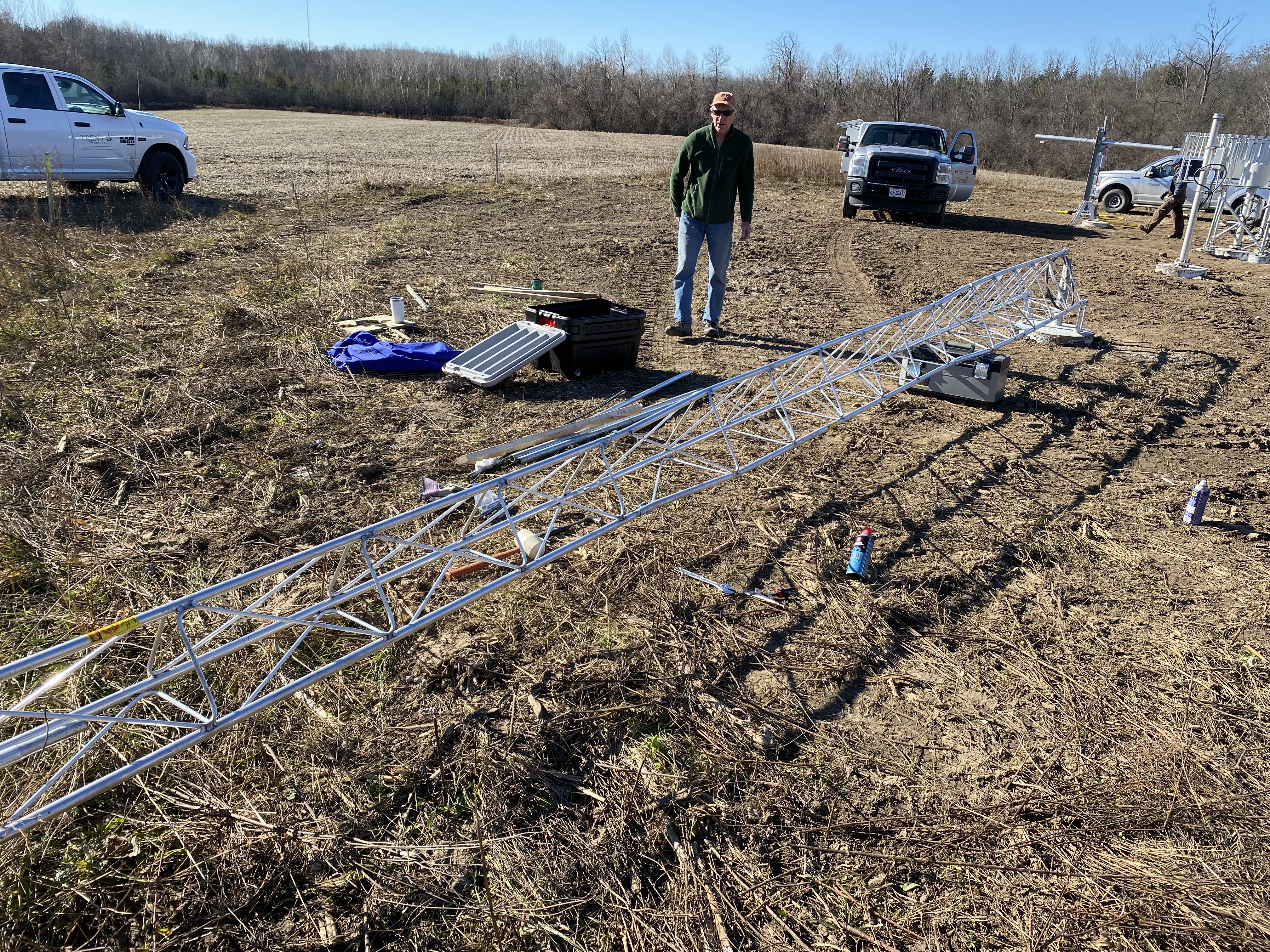 white metal tower on brown dirt field; blue sky and brown trees in background; one white man wearing beige cap, green jacket and blue jeans; three white trucks in background