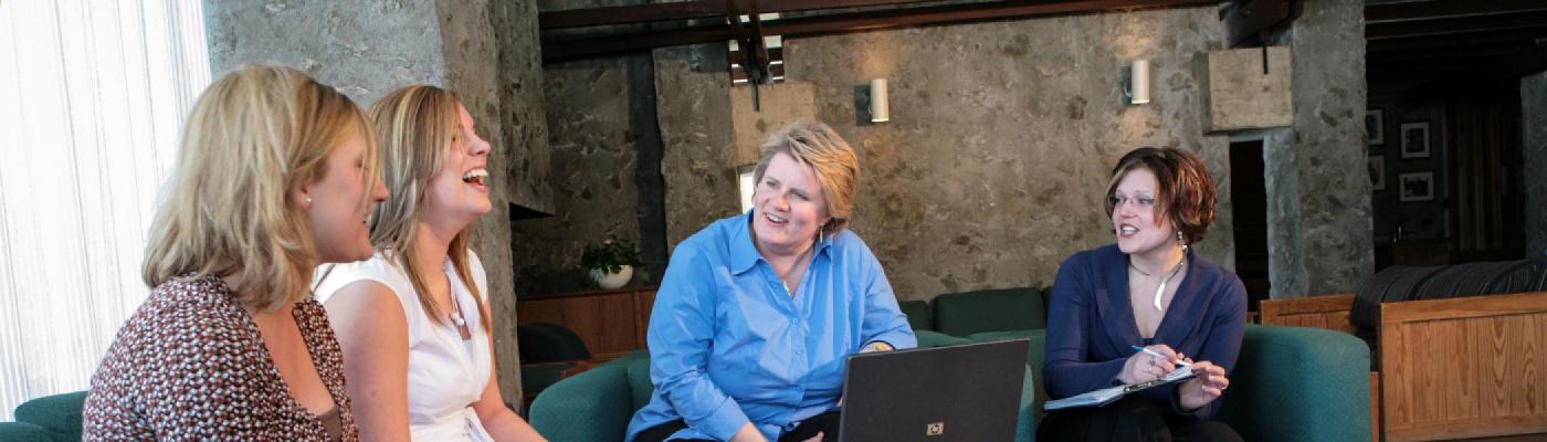 4 women sitting at a round table talking and smiling, in front of a laptop and books