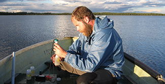 A man sitting in a boat on a blue lake taking awater sample, in a blue rain coat