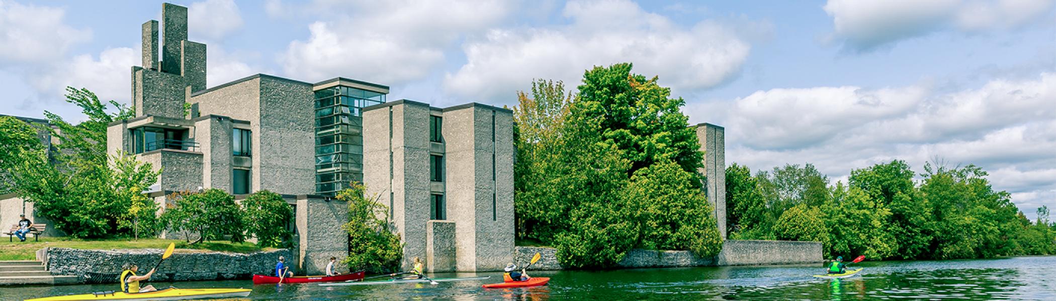 Four people kayaking downstream in front of Champlain college on a sunny day.