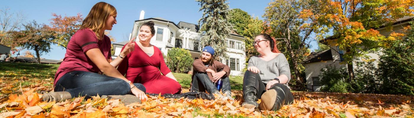 Four students sitting in a pile of leaves outside Traill College