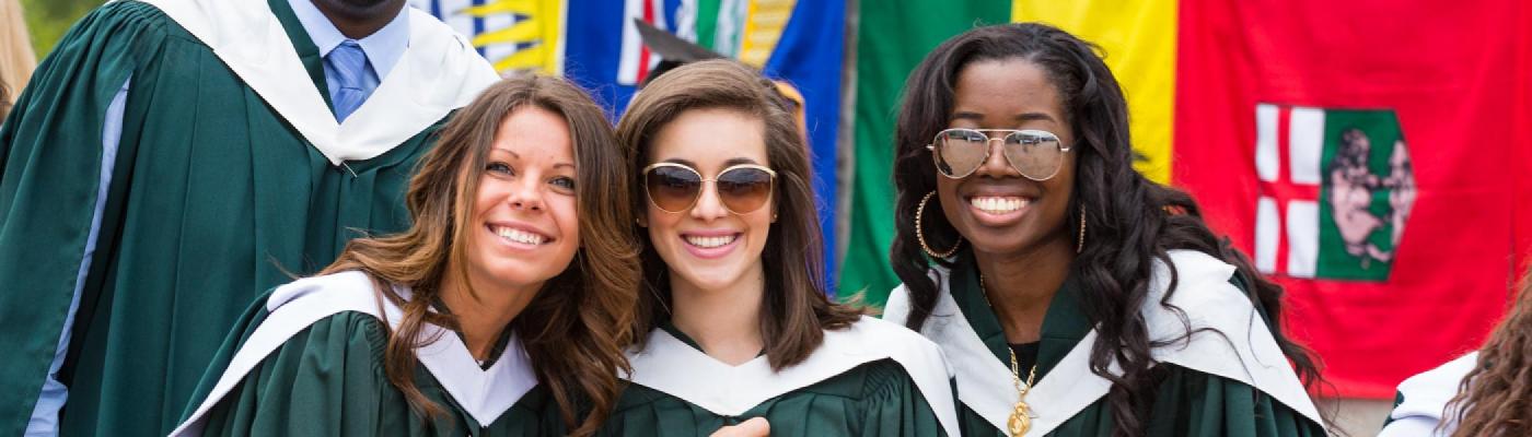 3 History students dressed in convocation gowns standing in front of the international flags, smiling at the camera