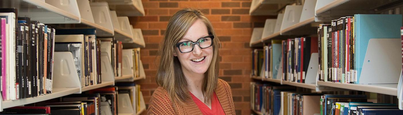 A female student standing in the library, in between two bookcase holding a book and smiling at the camera