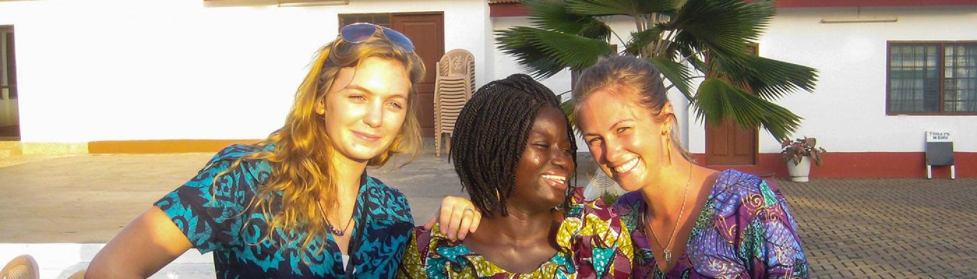 3 International Development Studies students smiling at the camera, in a Ghana yard in the sun