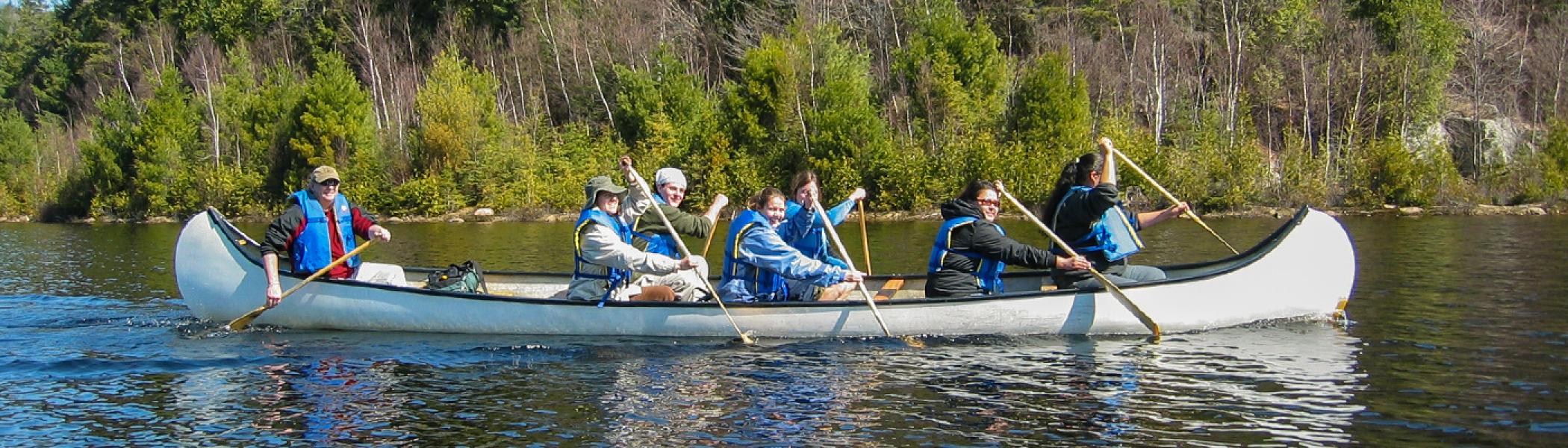 a group of people sitting in a canoe with blue life jackets on, paddling along a river in the summer sun